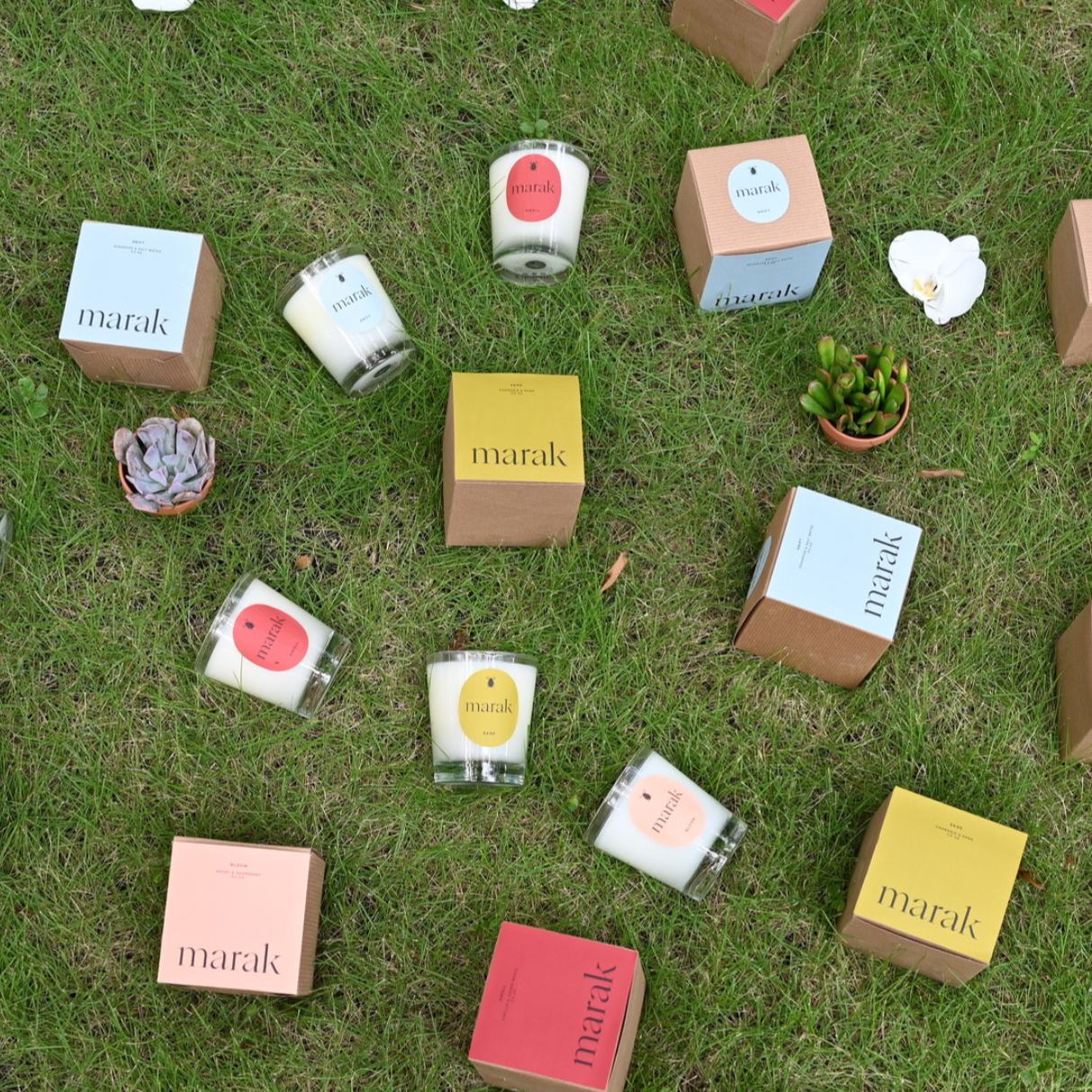 Various 'marak' products including candles and boxes scattered on grass with flowers.